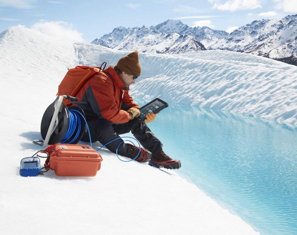 Man wearing sunglasses, protective jacket, gloves and cramp-on ice climbing shoes seated on a glacier holding a Dell Latitude 12 Rugged Tablet (Model 7202) computer. <a href=